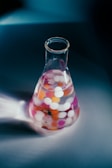 A glass Erlenmeyer flask containing a clear liquid is filled with multicolored spherical objects, possibly representing a science experiment. The flask sits on a smooth, reflective surface, casting a soft shadow.