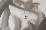 A delicate silver necklace with a circular pendant featuring intricate blue and white detailing is placed on a softly textured white fabric. The lighting creates subtle shadows and highlights, enhancing the elegance of the necklace and fabric.