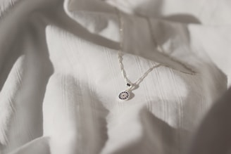 A delicate silver necklace with a circular pendant featuring intricate blue and white detailing is placed on a softly textured white fabric. The lighting creates subtle shadows and highlights, enhancing the elegance of the necklace and fabric.
