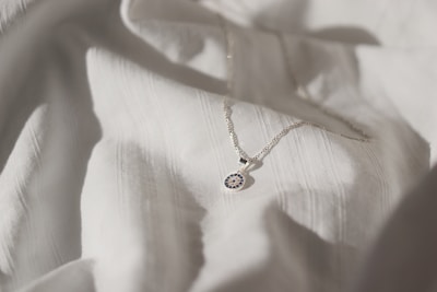 A delicate silver necklace with a circular pendant featuring intricate blue and white detailing is placed on a softly textured white fabric. The lighting creates subtle shadows and highlights, enhancing the elegance of the necklace and fabric.