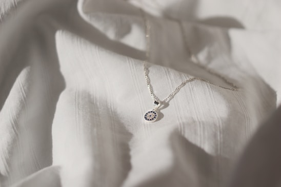 A delicate silver necklace with a circular pendant featuring intricate blue and white detailing is placed on a softly textured white fabric. The lighting creates subtle shadows and highlights, enhancing the elegance of the necklace and fabric.