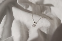 A delicate silver necklace with a circular pendant featuring intricate blue and white detailing is placed on a softly textured white fabric. The lighting creates subtle shadows and highlights, enhancing the elegance of the necklace and fabric.