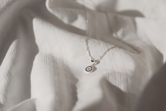 A delicate silver necklace with a circular pendant featuring intricate blue and white detailing is placed on a softly textured white fabric. The lighting creates subtle shadows and highlights, enhancing the elegance of the necklace and fabric.
