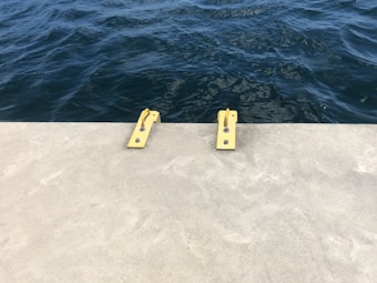 A concrete dock or pier is adjacent to dark blue rippling water. Two yellow metal bollards are secured to the dock, typically used for mooring boats.