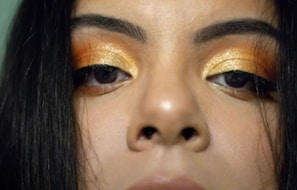 A close-up view of a person's eyes, showing glittering gold and bronze eye makeup. The eyebrows are thick and dark, and the hair frames the face on either side. The overall focus is on the intricately applied eye makeup.