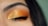 An eye with closed eyelid displaying vibrant, shimmering orange and gold eyeshadow. The eyelashes are long and well-defined. The eyebrow appears well-groomed, and the skin tone complements the warm hues of the makeup.