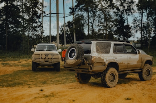 Two off-road vehicles covered in mud are parked on a dirt area with a backdrop of tall trees and a water tower in the distance. The vehicles appear to have been driven through challenging terrain, indicated by the heavy mud. The scene is outdoors, likely in a rural or forested area.