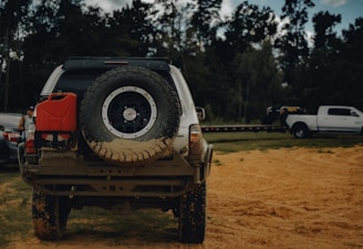 A rugged off-road vehicle with a large spare tire mounted on the back and a red fuel canister attached to the rear. The vehicle is parked on a sandy terrain, surrounded by trees. In the background, there is a white pickup truck with a trailer.