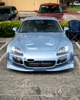 A silver sports car with a customized body kit is parked in an open parking lot. The car has distinctive front vents and headlights, and its windows are tinted. There's a red SUV and a black sedan parked behind it, along with a folded chair placed on the sidewalk. Yellow flowers and greenery are visible in the background.