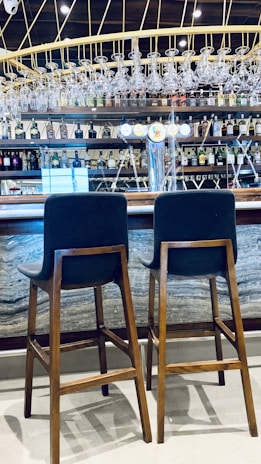 Two wooden bar stools with dark cushions are placed in front of a bar counter. The bar features a display of various liquor bottles neatly arranged on shelves and an overhead rack of clean glassware. The setting includes bright lighting and a polished countertop, creating a modern and inviting atmosphere.