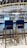 Two wooden bar stools with dark cushions are placed in front of a bar counter. The bar features a display of various liquor bottles neatly arranged on shelves and an overhead rack of clean glassware. The setting includes bright lighting and a polished countertop, creating a modern and inviting atmosphere.
