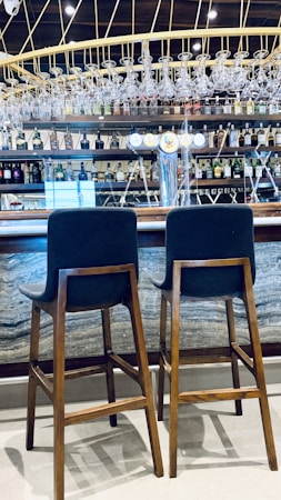 Two wooden bar stools with dark cushions are placed in front of a bar counter. The bar features a display of various liquor bottles neatly arranged on shelves and an overhead rack of clean glassware. The setting includes bright lighting and a polished countertop, creating a modern and inviting atmosphere.