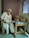 An elderly man sits on a chair beside a bed, holding the hand of a person lying down. The setting is a simple room with plain walls and a window. An intravenous drip is visible, indicating a healthcare environment.