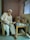 An elderly man sits on a chair beside a bed, holding the hand of a person lying down. The setting is a simple room with plain walls and a window. An intravenous drip is visible, indicating a healthcare environment.