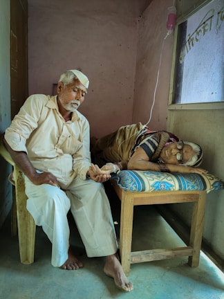 An elderly man sits on a chair beside a bed, holding the hand of a person lying down. The setting is a simple room with plain walls and a window. An intravenous drip is visible, indicating a healthcare environment.