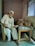 An elderly man sits on a chair beside a bed, holding the hand of a person lying down. The setting is a simple room with plain walls and a window. An intravenous drip is visible, indicating a healthcare environment.