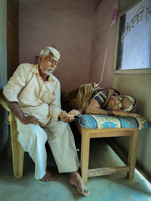 An elderly man sits on a chair beside a bed, holding the hand of a person lying down. The setting is a simple room with plain walls and a window. An intravenous drip is visible, indicating a healthcare environment.