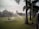 A large green lawn is being watered by sprinklers, with palm trees lining the right side and a pathway visible. In the background, there is a building with a dome and a tall water tower. The sky appears slightly overcast, adding to the serene setting.