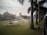 A large green lawn is being watered by sprinklers, with palm trees lining the right side and a pathway visible. In the background, there is a building with a dome and a tall water tower. The sky appears slightly overcast, adding to the serene setting.