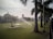 A large green lawn is being watered by sprinklers, with palm trees lining the right side and a pathway visible. In the background, there is a building with a dome and a tall water tower. The sky appears slightly overcast, adding to the serene setting.