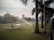 A large green lawn is being watered by sprinklers, with palm trees lining the right side and a pathway visible. In the background, there is a building with a dome and a tall water tower. The sky appears slightly overcast, adding to the serene setting.