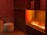A room with warm lighting features a modern fireplace with visible flames. In the foreground, there's a round houndstooth-patterned pouf next to a dark wooden side table. The wall and shelving unit is composed of dark wood, adding to the cozy and intimate ambiance.