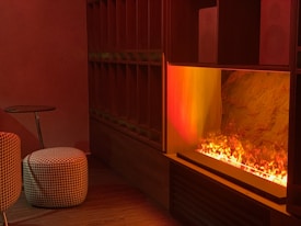 A room with warm lighting features a modern fireplace with visible flames. In the foreground, there's a round houndstooth-patterned pouf next to a dark wooden side table. The wall and shelving unit is composed of dark wood, adding to the cozy and intimate ambiance.