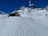 A snowy mountain landscape features a chairlift in motion and a small lodge with a stone exterior. The snowy foreground is marked with ski tracks, and a few people in bright clothing are gathered near the building. Rugged snow-covered peaks rise against a cloudless blue sky.