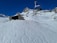 A snowy mountain landscape features a chairlift in motion and a small lodge with a stone exterior. The snowy foreground is marked with ski tracks, and a few people in bright clothing are gathered near the building. Rugged snow-covered peaks rise against a cloudless blue sky.