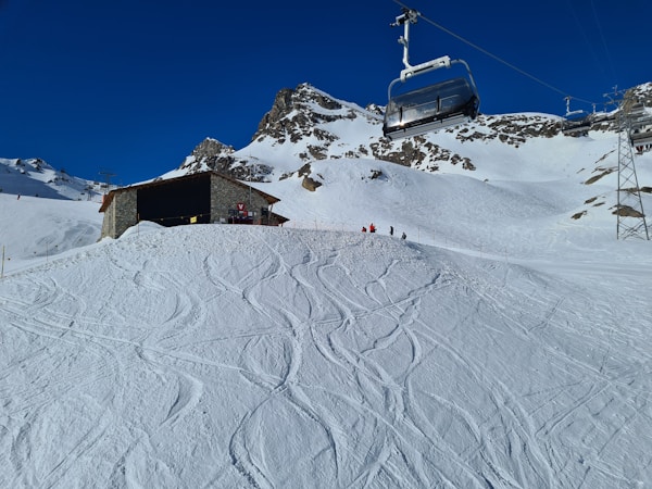 A snowy mountain landscape features a chairlift in motion and a small lodge with a stone exterior. The snowy foreground is marked with ski tracks, and a few people in bright clothing are gathered near the building. Rugged snow-covered peaks rise against a cloudless blue sky.