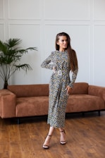 A woman stands confidently in a stylish floral dress with long sleeves and a belted waist, her long hair flowing over her shoulders. She is posed in a modern living room with hardwood floors, a brown suede couch, and a tall potted plant in the background. The wall is decorated with minimalist paneling.