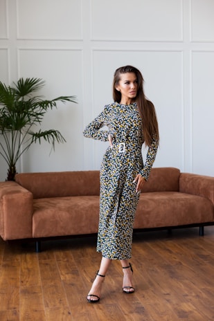 A woman stands confidently in a stylish floral dress with long sleeves and a belted waist, her long hair flowing over her shoulders. She is posed in a modern living room with hardwood floors, a brown suede couch, and a tall potted plant in the background. The wall is decorated with minimalist paneling.