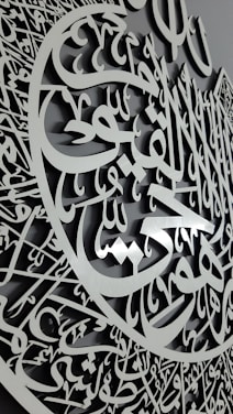 A detailed carving featuring intricate Arabic calligraphy with an elegant script and a three-dimensional appearance. The calligraphy is arranged in a circular pattern, suggesting a decorative and artistic design.