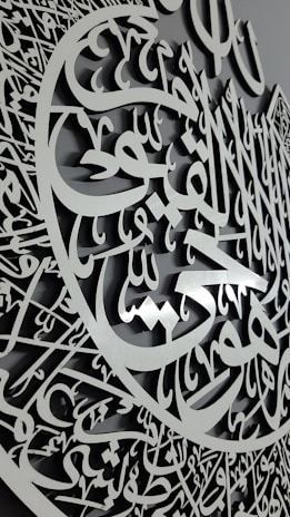 A detailed carving featuring intricate Arabic calligraphy with an elegant script and a three-dimensional appearance. The calligraphy is arranged in a circular pattern, suggesting a decorative and artistic design.