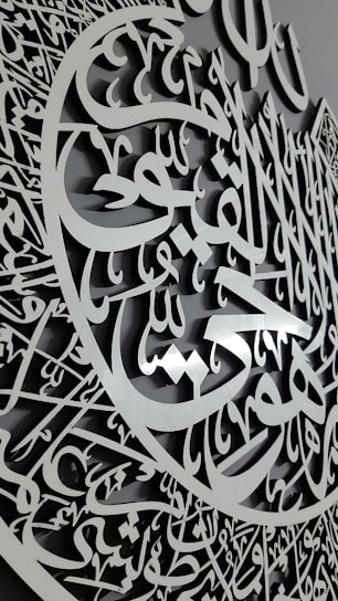 A detailed carving featuring intricate Arabic calligraphy with an elegant script and a three-dimensional appearance. The calligraphy is arranged in a circular pattern, suggesting a decorative and artistic design.