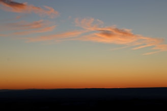 A serene landscape featuring a vast sky painted with soft pastel hues of orange and pink. Light, wispy clouds stretch across the horizon, with the subtle transition of colors indicating a setting or rising sun. The horizon itself is a dark, shadowed silhouette, adding contrast to the vibrant sky above.