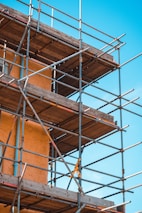 A construction scaffold attached to a building under a clear blue sky. The scaffolding is made of metal tubes and wooden planks, surrounding a building with an orange stucco finish. Yellow safety tape is visible on some parts of the structure.