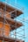 A construction scaffold attached to a building under a clear blue sky. The scaffolding is made of metal tubes and wooden planks, surrounding a building with an orange stucco finish. Yellow safety tape is visible on some parts of the structure.