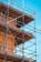 A construction scaffold attached to a building under a clear blue sky. The scaffolding is made of metal tubes and wooden planks, surrounding a building with an orange stucco finish. Yellow safety tape is visible on some parts of the structure.