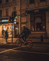 A cyclist with a backpack is riding a bike on a city street at night. The street is lit by warm streetlights, and there is a lit building with signage in the background. A traffic light is visible, displaying green. Another pedestrian and cyclist are also present in the scene.