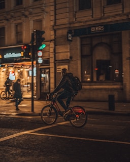 A cyclist with a backpack is riding a bike on a city street at night. The street is lit by warm streetlights, and there is a lit building with signage in the background. A traffic light is visible, displaying green. Another pedestrian and cyclist are also present in the scene.