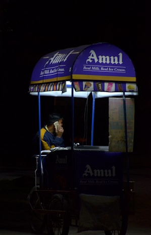 A person is sitting inside a small mobile ice cream cart with the Amul logo prominently displayed. The cart is primarily blue and purple with illuminated signs, and the person appears to be on a phone call. The setting seems to be dark, possibly at night, with the cart lit up against the surrounding darkness.