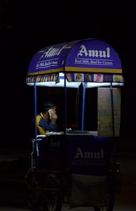 A person is sitting inside a small mobile ice cream cart with the Amul logo prominently displayed. The cart is primarily blue and purple with illuminated signs, and the person appears to be on a phone call. The setting seems to be dark, possibly at night, with the cart lit up against the surrounding darkness.