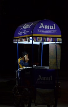 A person is sitting inside a small mobile ice cream cart with the Amul logo prominently displayed. The cart is primarily blue and purple with illuminated signs, and the person appears to be on a phone call. The setting seems to be dark, possibly at night, with the cart lit up against the surrounding darkness.