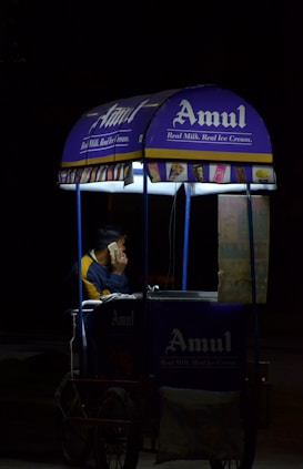 A person is sitting inside a small mobile ice cream cart with the Amul logo prominently displayed. The cart is primarily blue and purple with illuminated signs, and the person appears to be on a phone call. The setting seems to be dark, possibly at night, with the cart lit up against the surrounding darkness.