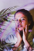 A person is surrounded by vibrant, multicolored light effects, with their hands gently placed on their face. Green leaves partially frame the subject, adding an element of nature to the scene.