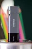 A Samsung USB drive is standing upright on a reflective surface. The device is predominantly gray with a black base that features a small 'USB 3.1' label. In the background, colorful blurred objects, likely pencils or crayons, create a vibrant contrast against a dark green backdrop.