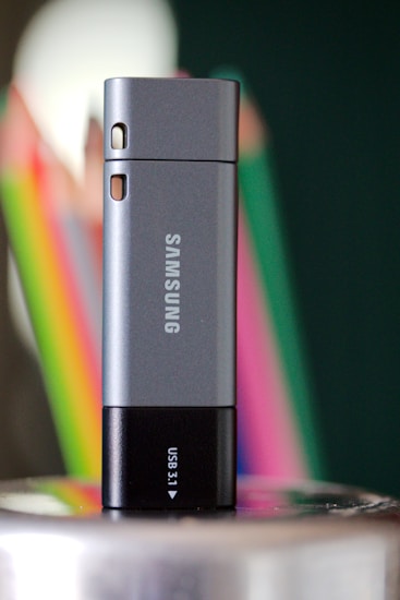 A Samsung USB drive is standing upright on a reflective surface. The device is predominantly gray with a black base that features a small 'USB 3.1' label. In the background, colorful blurred objects, likely pencils or crayons, create a vibrant contrast against a dark green backdrop.