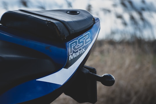 A close-up view of the back seat of a motorcycle with the label 'RS2 Matrix' in bold lettering. The bike seat is black with blue and white details surrounding the logo. The background is blurred, indicating an outdoor setting possibly with grass or vegetation.