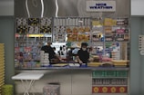 A small retail kiosk is filled with a variety of packaged food items like snacks, cereal boxes, and candy. Behind the counter, a person wearing a mask is organizing or checking inventory while standing in front of a display with a 'NICE WEATHER' sign above. The countertop has stacks of trays and a few other items.